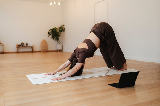 Woman holds downward dog yoga pose on a mat in a warm home studio, calm wellness style against a light interior background