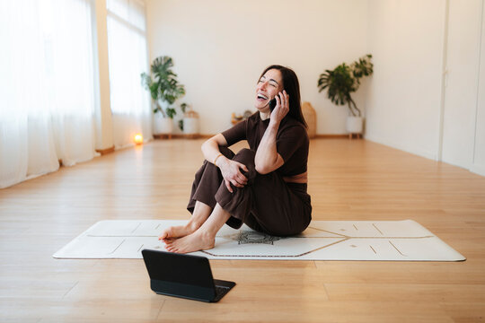 Joyful woman laughing on a phone during home yoga practice, lifestyle photography, bright wooden studio background, wellness and connection