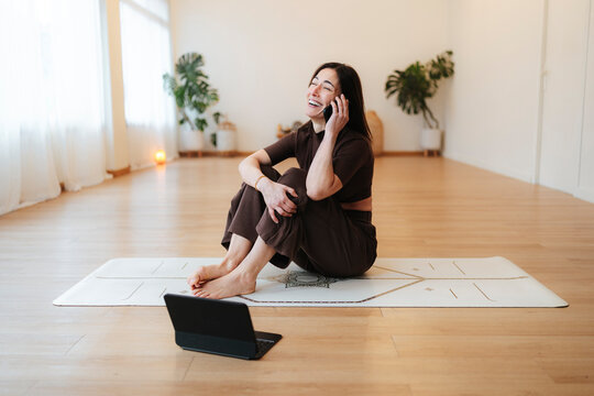 Smiling woman talks on a smartphone while seated on a yoga mat in a bright minimalist room, lifestyle photo about mindful balance