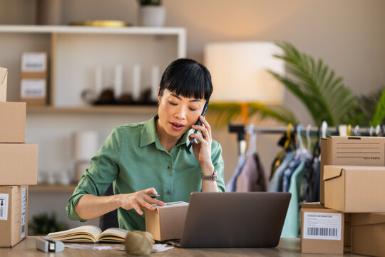 Woman packing orders for small online clothing business while managing customer calls and laptop