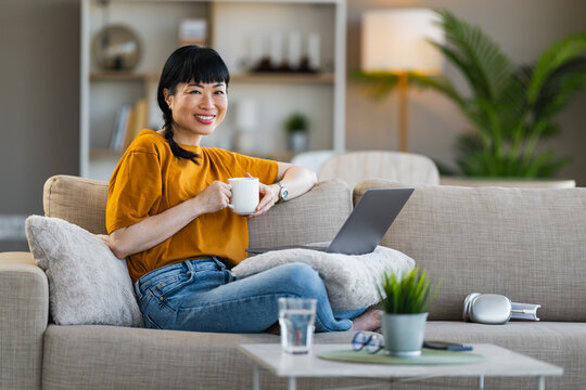 Smiling woman working from home on laptop while relaxing on sofa with a cup of coffee