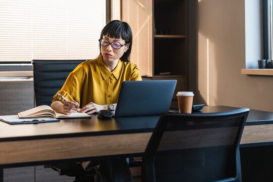 Businesswoman working at desk in modern office writing notes next to laptop and coffee