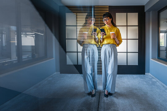 Young professional woman in office checking smartphone while holding coffee and reflected in glass