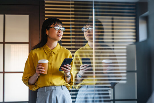 Young professional woman with coffee and smartphone looking through office glass reflected in window