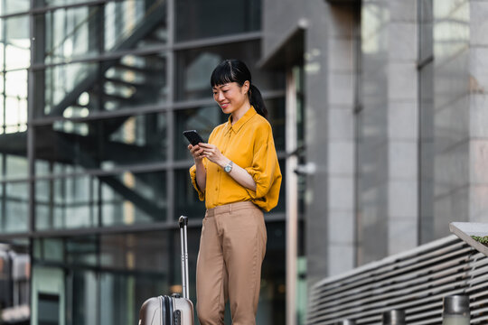Business traveler checks phone while waiting with suitcase outside modern office building