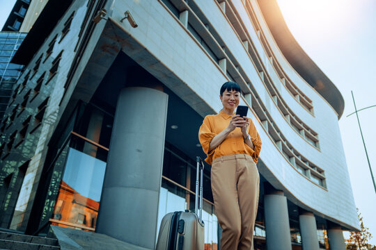 Business traveler woman using smartphone with rolling suitcase outside modern office building