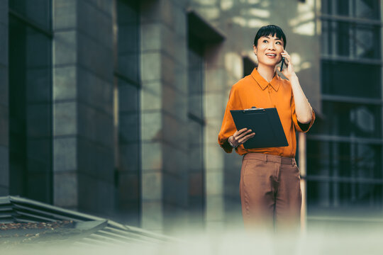 Young professional woman talking on phone while holding clipboard outside modern office building