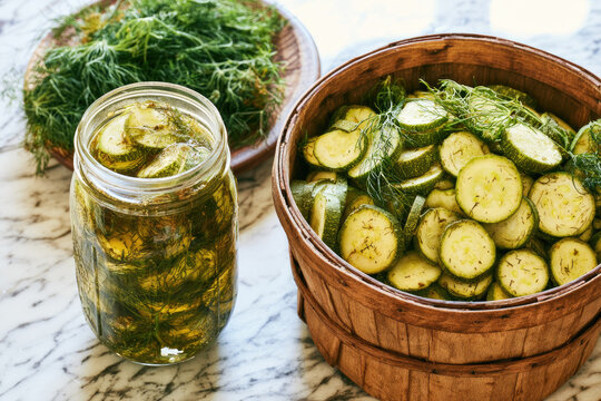 Pickled squash in glass jar beside wooden basket with fresh dill on marble surface, sustainable food preservation and harvest surplus concept with bright natural light and copy space