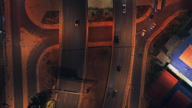 Dramatic top-down drone shot showing cars moving on a multi-lane highway at night, revealing an illuminated tennis court and complex road interchanges with warm streetlights