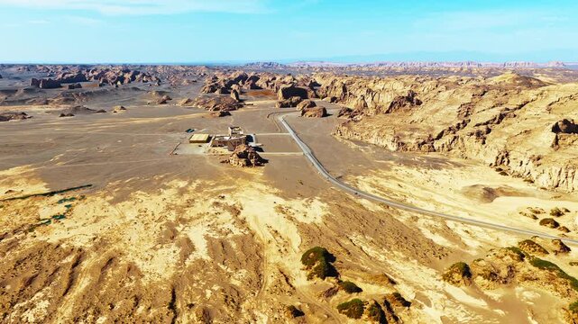Barren yardang desert landscape in Xinjiang, China, featuring eroded canyon cliffs, a winding road, sparse vegetation, and a remote facility under blue sky.
