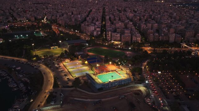 Stunning aerial footage of a brightly lit sports complex with a swimming pool and tennis courts, nestled within a coastal city of Athens, Greece as traffic moves along the surrounding roads at night