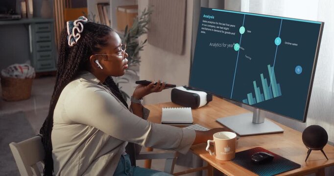 Focused young Black woman uses an AI assistant on her phone while working at a computer with business analytics visible on the screen in her living room.
