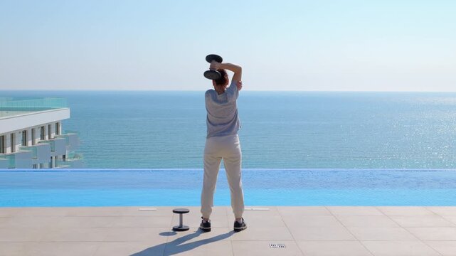 Woman doing triceps extension dumbbell exercise overlooking the sea. Back view of a young athletic woman training her arms and doing overhead dumbbell triceps extensions by an infinity pool