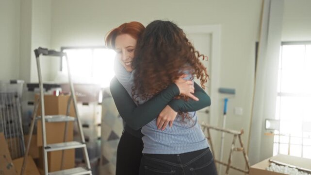 Two women hugging with arms around each other's backs among moving boxes in an apartment building; friendship joy.