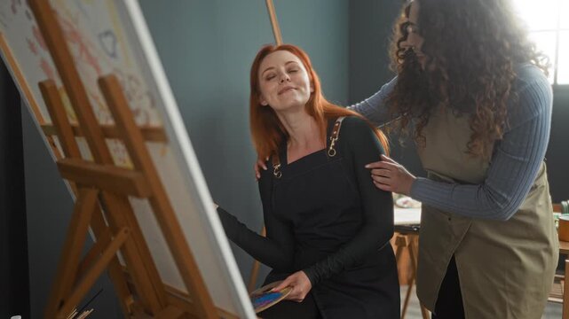 Woman painting at studio easel holding palette and brush while friend leans over with hand on shoulder, both smiling in apron clad studio workshop; creative collaboration joy.