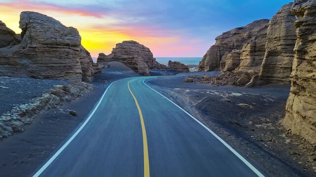 Winding paved road through an eroded rock canyon in Xinjiang, China, framed by layered cliffs and a colorful sunset sky, suited to travel and infrastructure themes.