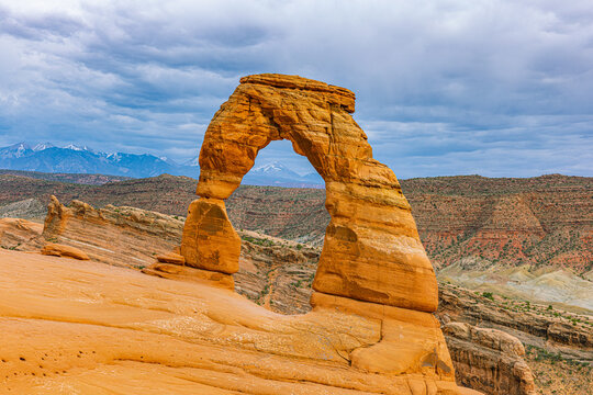 Famous Delicate Arch sandstone natural bridge standing against a dramatic cloudy sky with snowy peaks in the background at Arches National Park.