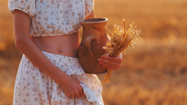 Woman in floral dress holding wheat and clay jug in golden hour field