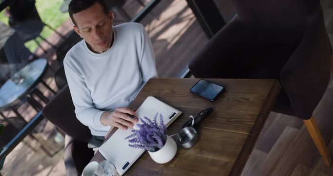 A man dressed in a casual sweater sketches on a notepad while seated at a wooden table in a bright and airy cafe. Lavender decor adds a touch of warmth to the setting