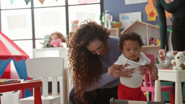 Woman supports toddler boy on wooden ride on toy in a colorful daycare playroom building; tenderness bonding.