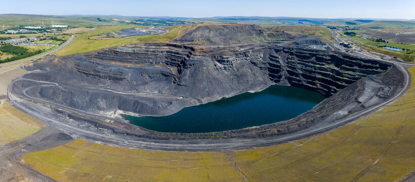 High angle panoramic view over closed opencast coal mine pit, dark water gathered at bottom of terraced rock face, former mining site pending land restoration