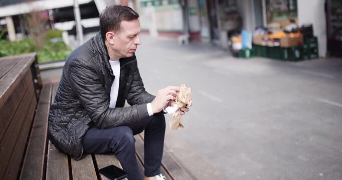 A man sits on a wooden bench in a lively city space, savoring tasty street food. Surrounding shops contribute to the vibrant atmosphere, making it an enjoyable moment