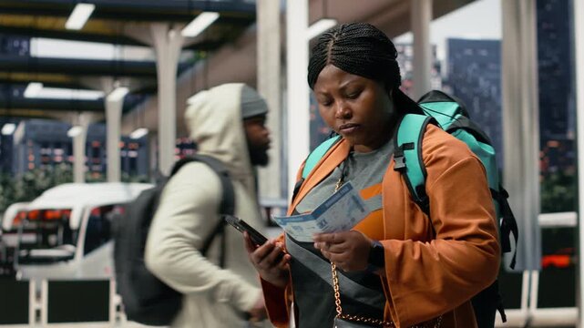 Close-up smiling woman comparing a printed bus ticket with information on her smartphone, checking travel document and route details on the coach platform before departure.