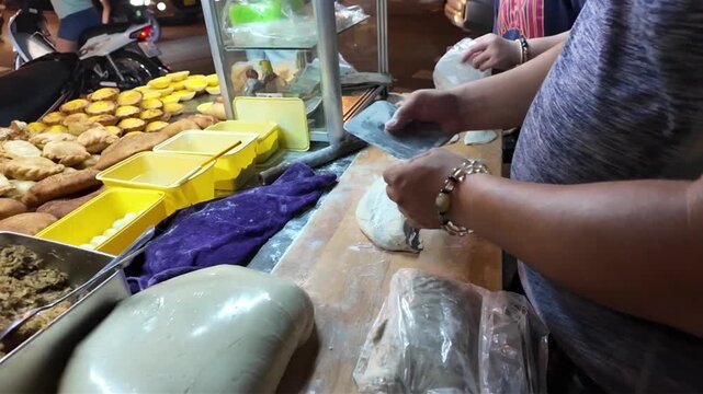 Close up video of a street food vendor kneading, cutting, and rolling dough on a wooden board to make crispy deep fried pork buns (banh bao chien) at a night food stall in Ho Chi Minh City, Vietnam