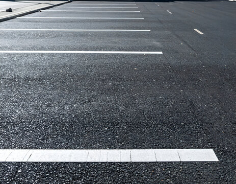 An empty asphalt parking lot with white painted parallel parking space markings. The surface is textured black pavement with crisp lines under bright daylight.