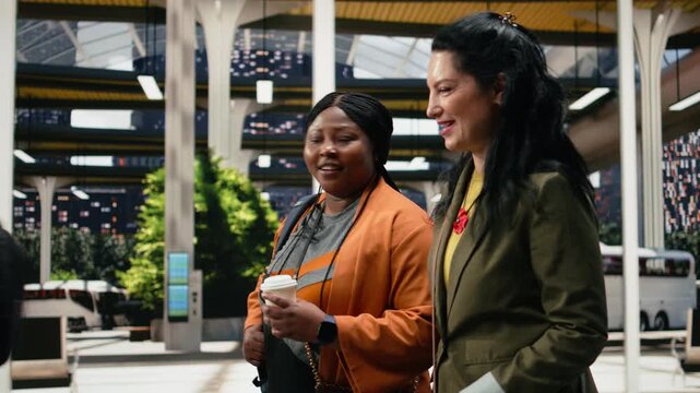 Cheerful women chat while crossing a coach station platform with coffee drinks; the friends carry takeaway cups and discuss their departure plans.
