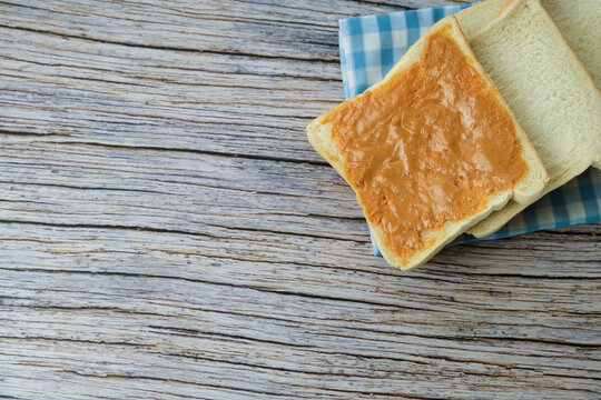Soft bread slices rest on a checkered cloth. Wooden table shows weathered, natural grain. One slice topped with ritz, peanut jam. Simple, cozy, inviting breakfast scene. Ideal for morning meal, cafe