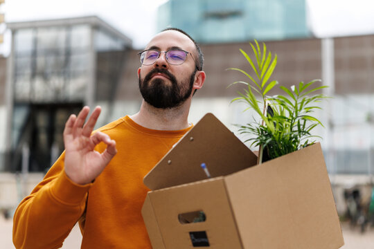 Young man holding a moving box with a plant and clipboard, making an OK gesture during an office relocation and starting a new job