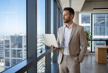 Fototapeta premium Businessman holding laptop looking out window in modern office
