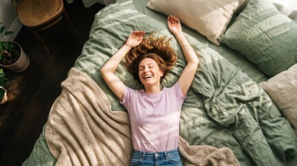Fototapeta premium An overhead view portrait of a happy young woman laughing with eyes closed and arms raised, lying on a cozy bed with green linen in a sunlit bedroom.