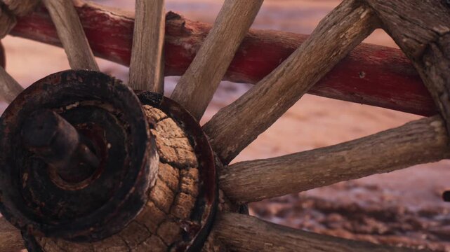 A wooden wheel of a cart is shown close up. It is resting on the ground in a rural area. The wood is aged and shows signs of wear. The background is simple and natural.