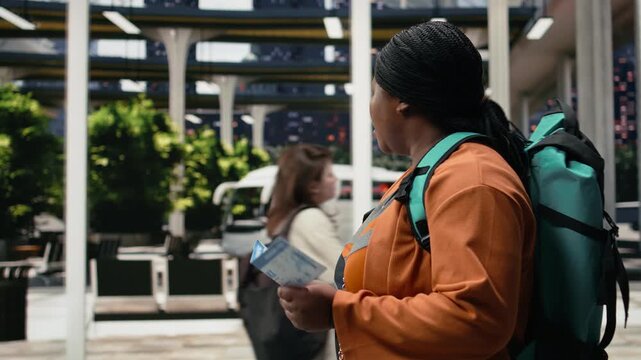 Woman reads a travel document and checks route details on a mobile phone inside a bus station; the traveler compares printed boarding information with smartphone updates before departure.