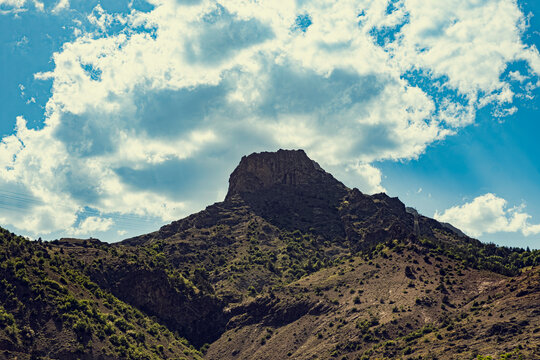 Rocky mountain peak. Flat summit rises above scrub-covered slopes against a blue sky with dramatic clouds. Ideal for travel, adventure, outdoor recreation, and nature backgrounds.