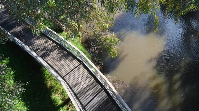 A high-angle, elevated shot looking down on a curving timber boardwalk path running alongside the Werribee River. Native eucalyptus branches frame the view of&nbsp;murky river water