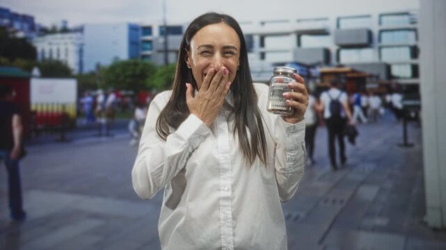 Hispanic woman holding a jar of money with hand over mouth gesture on busy street; surprise celebration.