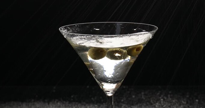 Fresh olives and water drops falling into glass with martini cocktail on black background, closeup