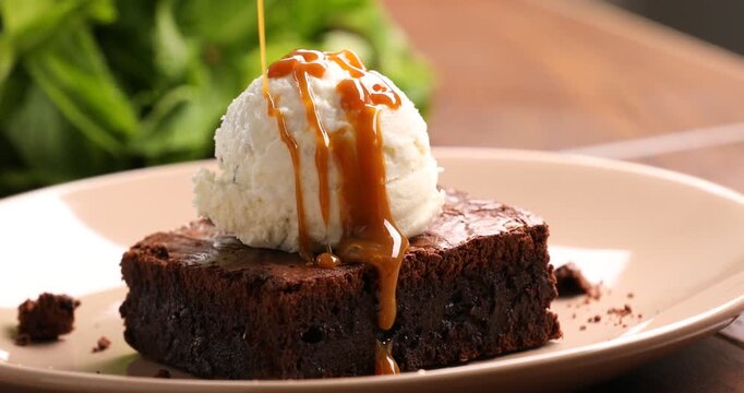 Pouring caramel topping over brownie with ice cream at wooden table, closeup