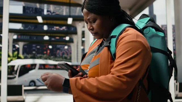 Passenger walks through a bus station with a phone and boarding pass; the solo traveler compares printed and digital details to confirm departure time.