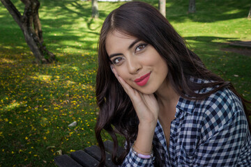 Fototapeta premium Close-up portrait of a beautiful young Latina woman smiling softly in a sunny city park. Outdoor lifestyle image with green background and natural daylight.