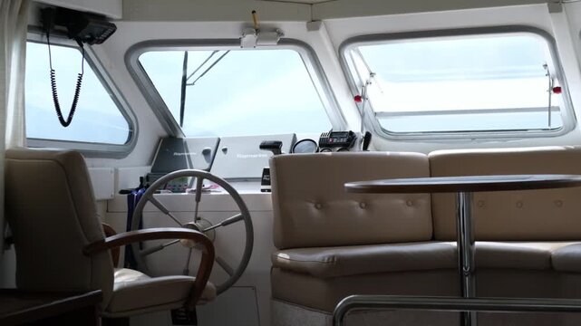 Interior view of a yacht's cabin showcasing steering wheel, seating area, and navigational equipment with large windows revealing a cloudy sky outside