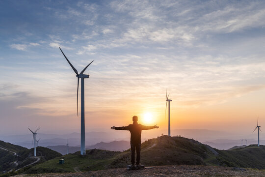 Man standing on mountain top with arms outstretched at sunset beside wind turbines