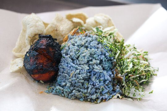 A delicious serving of Malaysian Nasi Kerabu featuring blue pea flower rice served with grilled chicken, raw vegetable and herb salad (ulam), and fish crackers.