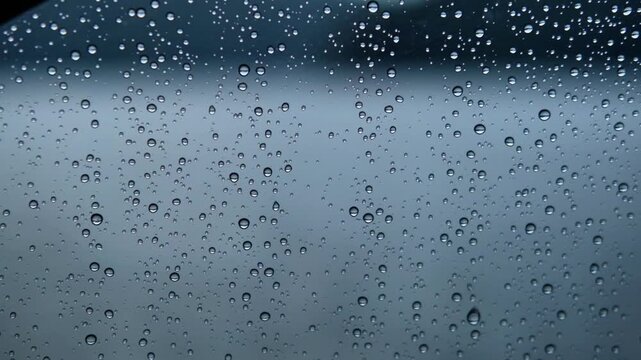 Close-up view of water droplets on a car window, showcasing the gradual accumulation and movement of raindrops against a blurred background of a rainy day