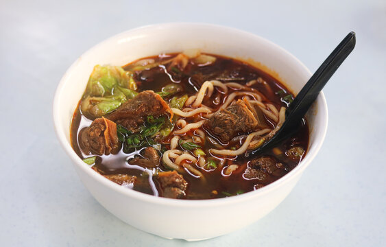 Braised beef noodle soup with tender beef chunks and greens in a rich spicy broth, served in a white bowl.