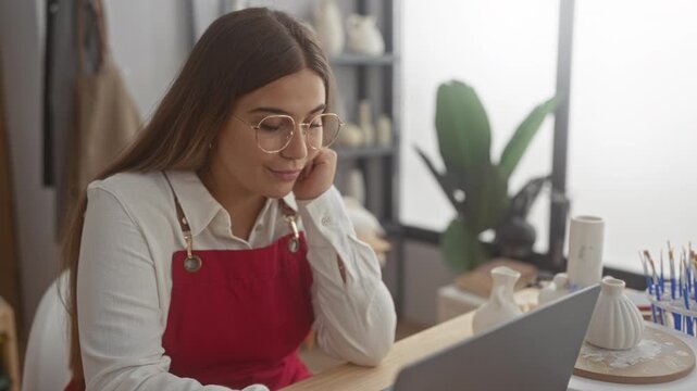 Woman in red apron rests chin on hand while studying a laptop and glazed pottery on a worktable in a ceramics studio; creative contemplation.