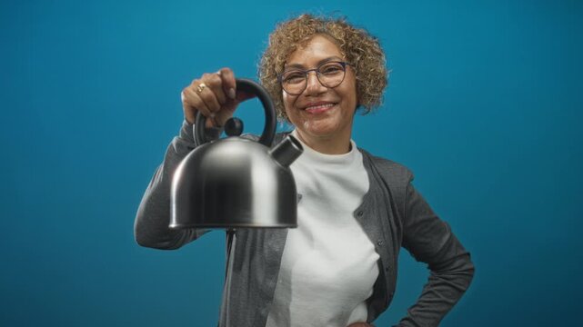 Middle age woman with curly hair and glasses holding a stainless steel kettle by the handle and smiling while wearing a cardigan in studio; cheerful.
